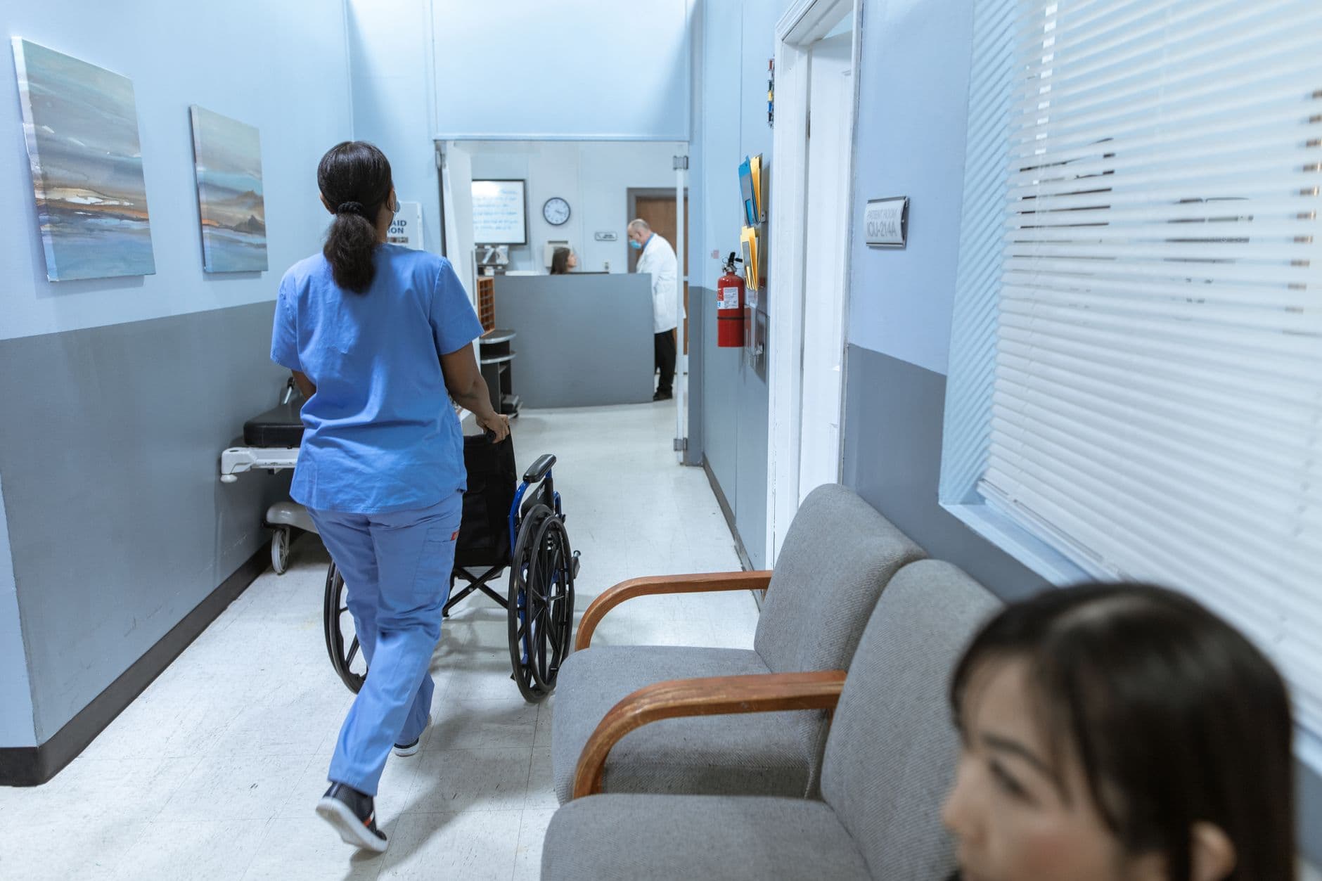 Clinic & Hospital Appointment Websites — A nurse in scrubs pushes a wheelchair through a well-lit hospital corridor, attending to patient care.
