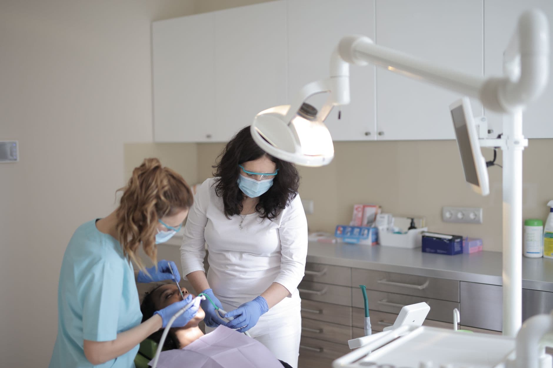 Dentist Appointment Websites — Young female dentist in medical mask and latex gloves curing teeth of patient with assistant using medical instruments in modern dental clinic