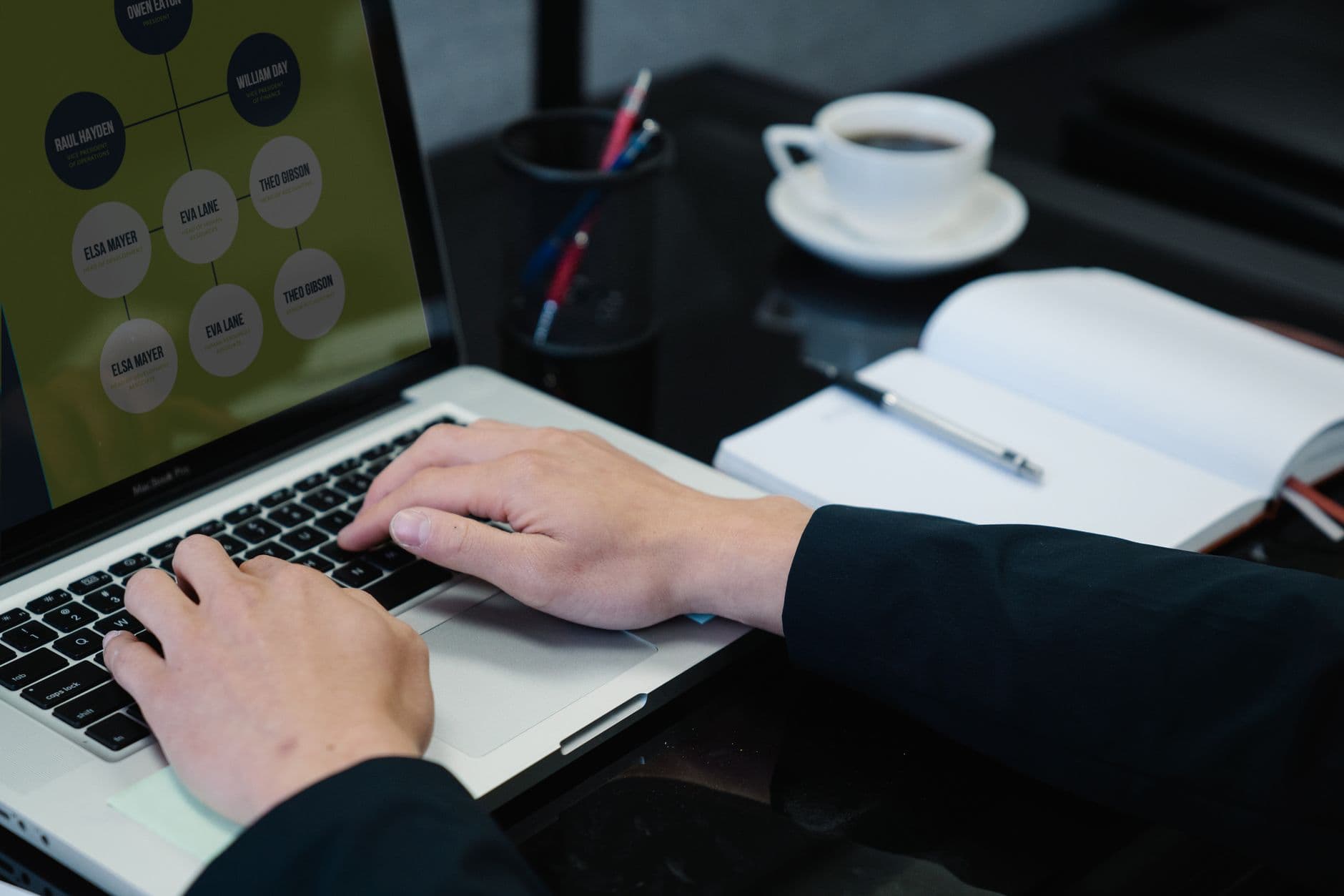 Business Automation — Close-up of hands typing on a laptop displaying an organizational chart, a notebook, and coffee nearby.