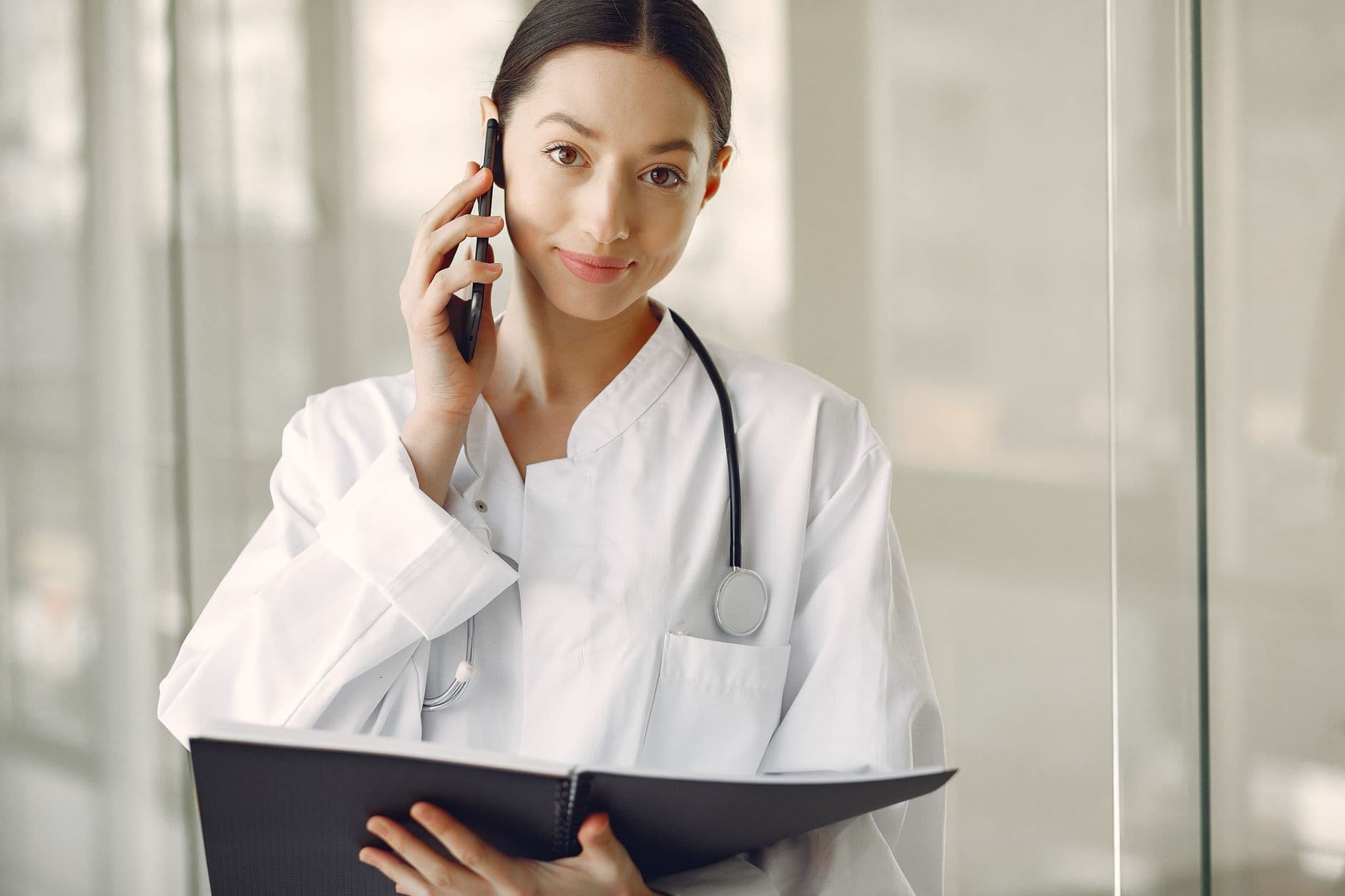 Website & Hosting for Doctors — Crop ethnic female doctor in medical uniform with stethoscope and notebook talking on cellphone while standing in clinic corridor